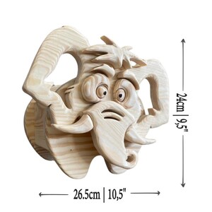 May include: A whimsical wooden sculpture of a cartoonish elephant head with large ears, tusks, and expressive eyes. The sculpture is 10.5 inches wide and 9.5 inches tall, crafted from light-colored wood.