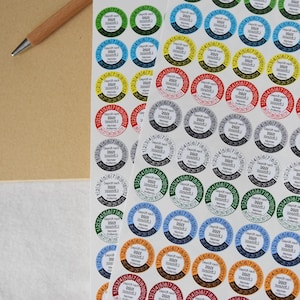 Könnte beinhalten: Blätter mit farbigen, kreisförmigen Aufklebern mit weißem Zentrum und Zahlenring. Die Aufkleber sind in verschiedenen Farben erhältlich, darunter Grün, Blau, Gelb, Rot und Orange. Ein Spiralnotizblock und ein Stift sind im Hintergrund.