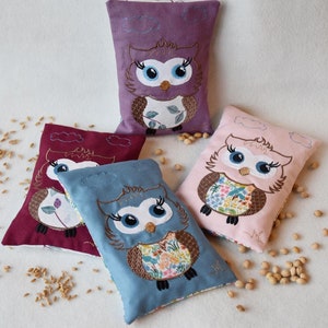 Könnte beinhalten: Vier Stoff-Wärmepads in Form einer Eule in verschiedenen Farben: Lila, Rot, Rosa und Blau. Jedes Pad hat ein anderes Blumenmuster und eine niedliche gestickte Eule mit großen Augen.