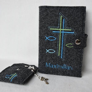 Könnte beinhalten: Ein dunkelgraues Filz-Bibelcover und ein kleines Täschchen-Set. Das Cover zeigt ein gesticktes Kreuz in Blau und Grün mit dem Namen "Maximilian" darunter. Das Set enthält einen Rosenkranz.