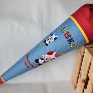 Könnte beinhalten: Eine blaue, rote und gelbe Schultüte mit dem Namen "THEO" in Gelb und Rot gestickt. Die Tüte zeigt gestickte Cartoonfiguren und den Text "Einschulung 2025". Die Tüte ist mit blauen und gelben Bändern gebunden.
