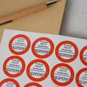 Könnte beinhalten: Blatt mit roten und weißen runden Aufklebern mit deutschem Text. Jeder Aufkleber trägt die Aufschrift "Prüfung NICHT bestanden GESPERRT". Ein Holzstift liegt auf einer braunen Oberfläche.