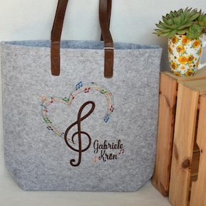 Könnte beinhalten: Graue Filz-Tragetasche mit braunen Ledergriffen. Die Tasche hat ein gesticktes herzförmiges Design mit Noten und einem Violinschlüssel. Der Name "Gabriele Krön" ist darunter gestickt.