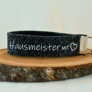 Könnte beinhalten: Ein grauer Filzschlüsselanhänger mit einem silbernen Ring und einem weißen gestickten Herz sowie dem Text "Hausmeister mit".