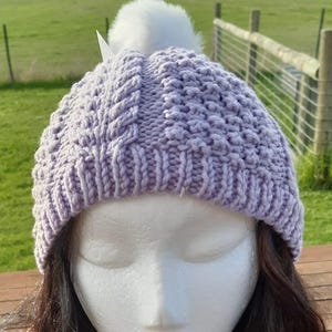 Könnte beinhalten: Eine lavendelfarbene Strickmütze mit einem weißen Kunstfell-Bommel. Die Mütze hat ein strukturiertes Strickmuster und einen gerippten Bund. Die Mütze wird auf einem Schaufensterkopf präsentiert.