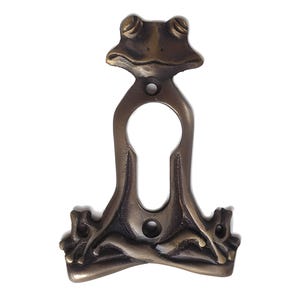 May include: A brass keyhole cover shaped like a frog sitting in a meditative pose. The frog has a detailed face and body, with a keyhole in the center.