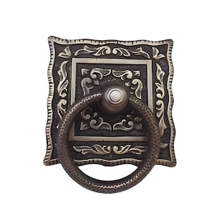 May include: A square-shaped, ornate, silver-toned door pull with a ring handle. The pull has a detailed, engraved design with a floral motif.