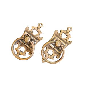 May include: Two ornate brass drawer pulls with a ring handle. The pulls have a detailed design with a crown-like top and intricate scrollwork.