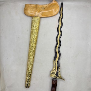 Op de afbeelding: Een keris-dolk met een golvend zwart lemmet met goudkleurige metalen inleg aan de basis, een donkerbruin houten handvat en een aparte goudkleurige metalen schede met gedetailleerde bloemgravures en een lichtgekleurde houten greep.