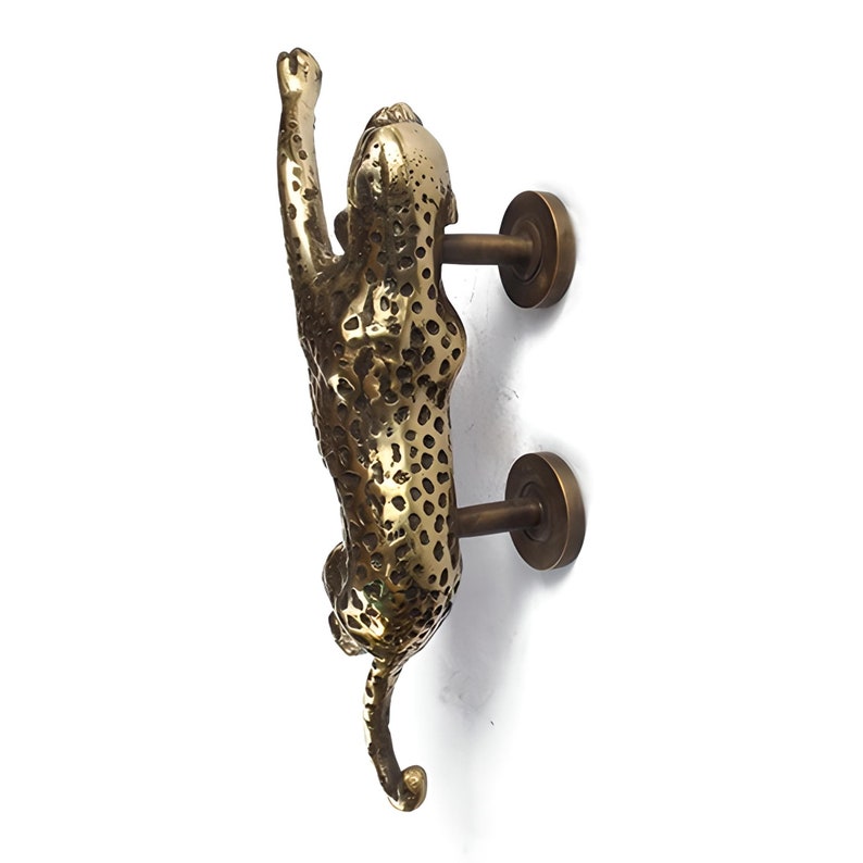 May include: A brass door handle shaped like a leopard reaching up. The handle is mounted on a white wall.