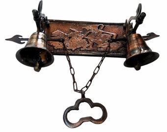 May include: A decorative wall-mounted double bell with a chain and a key-shaped pull. The bells are made of brass and have a copper finish. The plaque features a relief design of figures.