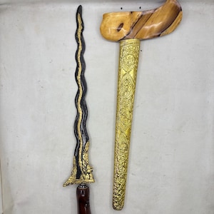 Op de afbeelding: Een keris-dolk met een golvend, donker metalen lemmet met goudkleurige accenten, een gepolijst donkerbruin houten handvat en een aparte goudkleurige metalen schede met ingewikkelde reliëfpatronen en een lichtbruin houten gevest.