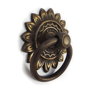 May include: A brass door knocker with a decorative sunflower design and a ring pull. The knocker has a detailed, ornate design with a circular centre and a ring that is attached to a round knob.