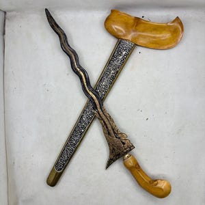 May include: A wavy-bladed dagger with a dark, textured metal blade and a light brown wooden handle. It is positioned across a long, slender scabbard featuring intricate silver-toned floral engravings and a wide, curved light brown wooden top.