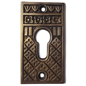 May include: A brass keyhole cover with an intricate geometric design. The cover features a square shape with a keyhole in the center and a decorative border.