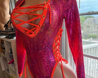 Könnte beinhalten: Ein langärmliger Body in einem leuchtenden Pink- und Orangeton. Der Body hat ein strukturiertes, reptilienartiges Muster und ist mit leuchtend orangefarbenen Zierleisten und Schnürdetails versehen. Das Design umfasst einen tiefen Ausschnitt und hoch geschnittene Beinausschnitte.