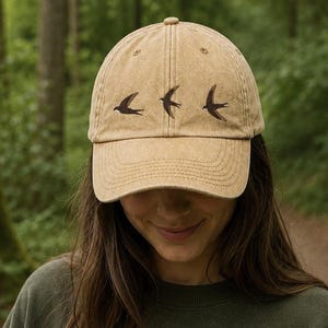 May include: A tan baseball cap with a distressed finish featuring three embroidered dark brown swallows across the front panels. The cap has a curved brim and a matching button on the top centre, showcasing a detailed bird motif.
