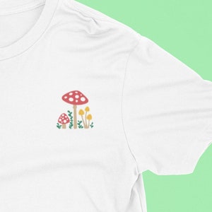 Könnte beinhalten: Weißes T-Shirt mit einem roten und weißen Pilz und gelben Pilzen, die auf der Vorderseite gestickt sind.