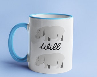 May include: White ceramic mug with a blue rim and handle. The mug features two gray hippopotamus illustrations and the word "will" in black cursive writing.