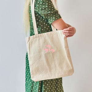 May include: A beige canvas tote bag featuring three pink shrimp illustrations. The bag has long straps and is held by a person wearing a green polka dot dress. The bag is a reusable shopping bag.