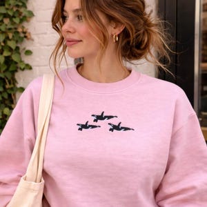 May include: A pale pink sweatshirt with a crew neck and long sleeves. The sweatshirt features a black embroidered design of three orcas. The model is wearing gold hoop earrings and has a cream-coloured tote bag.