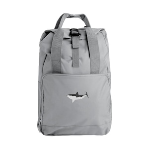 May include: A grey backpack featuring a shark design. The backpack has a top handle, adjustable straps, and a front pocket. The shark graphic is black, white, and grey. The backpack is made of a durable material and has a roll-top closure.
