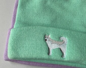 Peut inclure: Un bonnet vert avec un motif de chien brodé en blanc. Le bonnet a un bord rabattable violet.