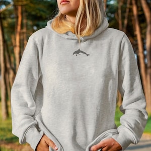 Puede incluir: Una sudadera con capucha gris con una pequeña silueta de delfín bordada en la parte delantera.