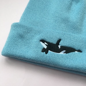 Op de afbeelding: Een lichtblauwe gebreide beanie met een geborduurd orka-ontwerp in zwart en wit.