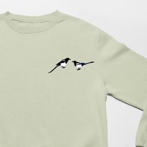 May include: A light green sweatshirt featuring an embroidered design of two magpies. The birds are depicted in black and white with blue accents. The sweatshirt has a crew neck and long sleeves.