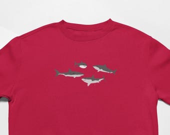 May include: A red sweatshirt featuring a graphic of four stylized sharks in shades of gray and white. The sharks are arranged in a playful composition, with two facing left and two facing right. The sweatshirt is made of a soft material.