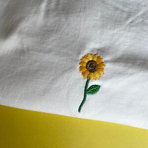 Puede incluir: Tela blanca con un girasol bordado. El girasol tiene p&eacute;talos amarillos, un centro marr&oacute;n, un tallo verde y una hoja verde. La tela est&aacute; sobre una superficie amarilla.