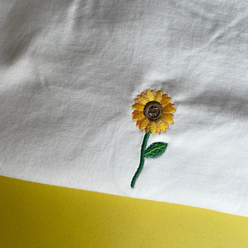 Puede incluir: Tela blanca con un girasol bordado. El girasol tiene p&eacute;talos amarillos, un centro marr&oacute;n, un tallo verde y una hoja verde. La tela est&aacute; sobre una superficie amarilla.