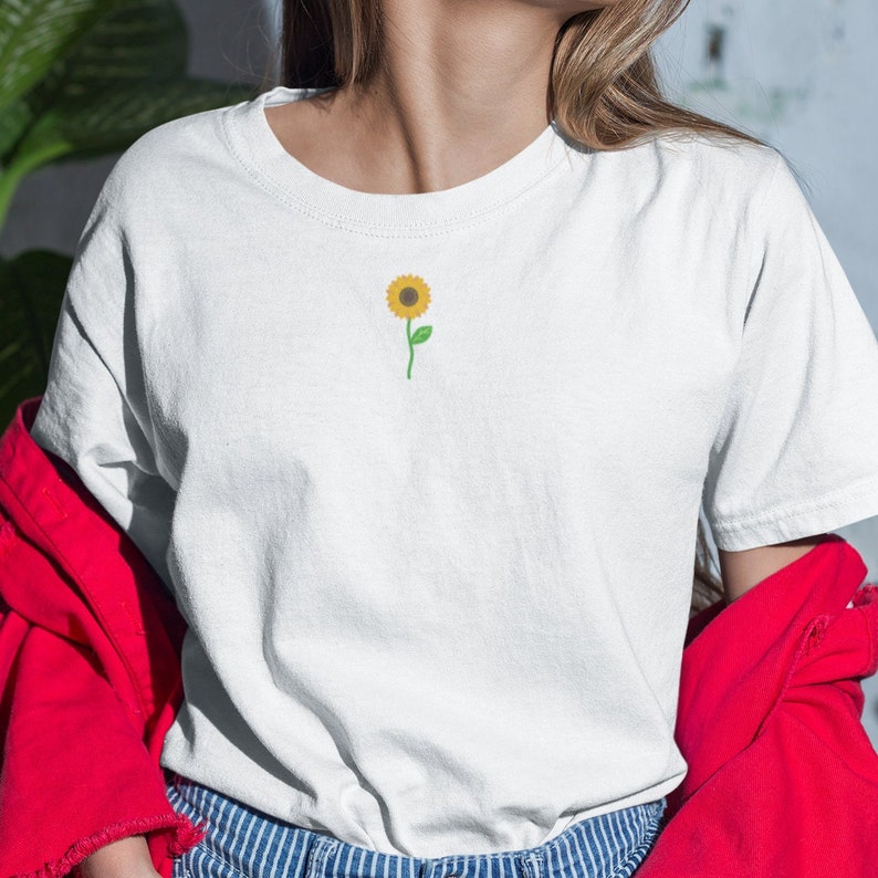 Puede incluir: Camiseta blanca con un peque&ntilde;o girasol bordado. El girasol tiene p&eacute;talos amarillos, un centro marr&oacute;n y un tallo y hojas verdes. La camiseta es de estilo cuello redondo cl&aacute;sico.