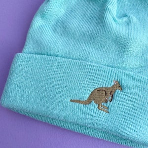 May include: A light blue beanie with an embroidered kangaroo design. The kangaroo is brown and has a joey in its pouch.