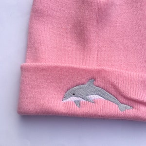 Puede incluir: Gorro de punto rosa con un delfín bordado gris en la parte delantera.