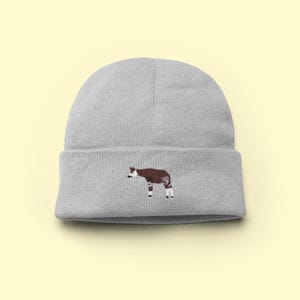 Può includere: Un berretto grigio chiaro con un ricamo di okapi. L'okapi è raffigurato in marrone e bianco, con un design dettagliato. Il berretto è realizzato in morbido materiale a maglia e ha un bordo ripiegato. Lo sfondo è giallo chiaro.