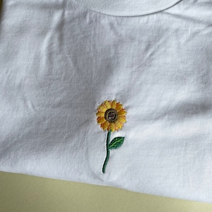 Puede incluir: Camiseta blanca con un peque&ntilde;o girasol bordado. El girasol tiene p&eacute;talos amarillos, un centro marr&oacute;n y un tallo verde con una sola hoja. La camiseta est&aacute; hecha de una tela blanca suave.
