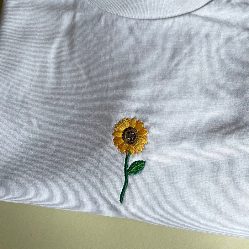 Puede incluir: Camiseta blanca con un peque&ntilde;o girasol bordado. El girasol tiene p&eacute;talos amarillos, un centro marr&oacute;n y un tallo verde con una sola hoja. La camiseta est&aacute; hecha de una tela blanca suave.