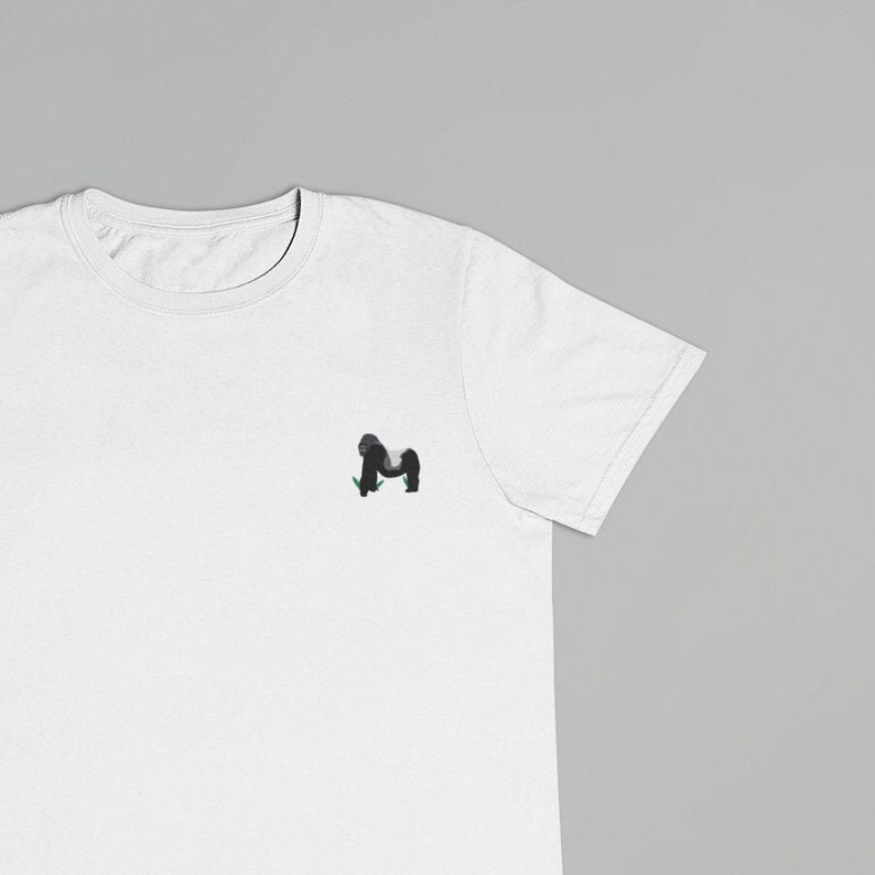 May include: A white t-shirt with a small black and gray gorilla graphic on the front. The gorilla is standing on a patch of green grass.