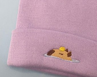 May include: A lavender knitted beanie with a small embroidered design of a brown and yellow animal with a yellow circle on its head.