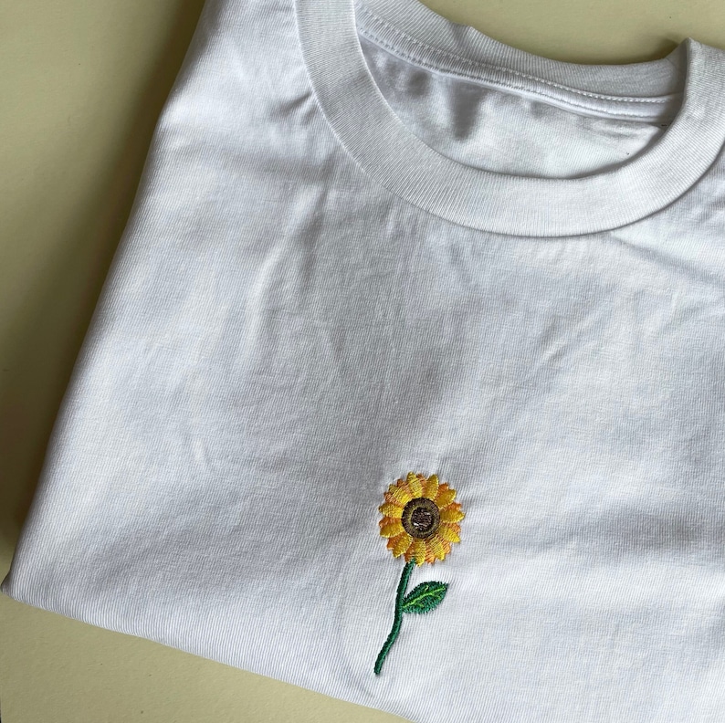 Puede incluir: Camiseta blanca con un peque&ntilde;o dise&ntilde;o de girasol bordado. El girasol tiene p&eacute;talos amarillos, un centro marr&oacute;n y un tallo verde con una sola hoja. La camiseta est&aacute; doblada.