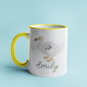 May include: A white ceramic mug with a yellow rim and handle. The mug features a cartoon illustration of a dodo bird and the name "Emily" written in cursive.