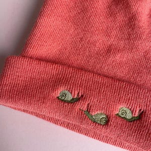 May include: A pink knitted beanie with three embroidered snails in green and brown.