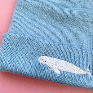 Pode incluir: Um gorro azul claro com um design de baleia beluga branca bordada.
