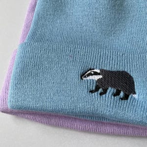 Könnte beinhalten: Eine blaue Strickmütze mit einem violetten Umschlag und einem weißen gestickten Dachs-Design.