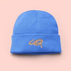 Pode incluir: Um gorro azul claro com um desenho de cobra castanha. O gorro é feito de um material macio e tricotado e apresenta uma aba dobrada. O desenho da cobra está na frente do gorro.