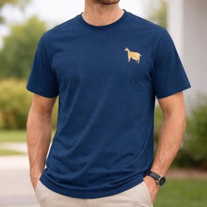 May include: Navy blue t-shirt with a gold goat graphic on the left chest. The short-sleeved shirt is made of a soft material and has a classic crew neck. The person is wearing khaki pants and a black watch.