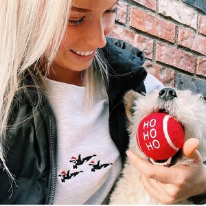 May include: A woman wearing a white long-sleeved shirt with a Christmas-themed design of penguins wearing Santa hats. She is holding a white dog with a red ball in its mouth that says "HO HO HO".