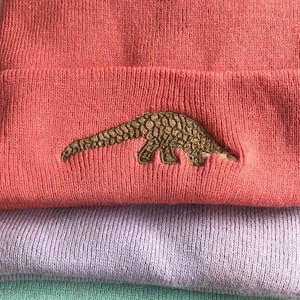 Puede incluir: Un gorro de color rosa coral con un diseño de pangolín bordado en oro.